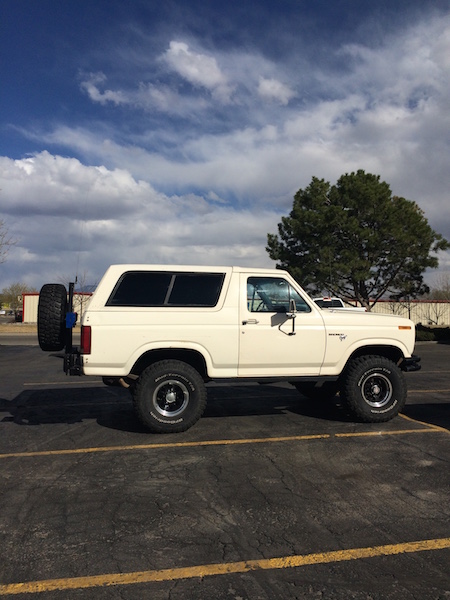 1981 Ford Bronco - A white 1980s Ford Bronco is parked in a lot under a partly cloudy sky. The SUV has a spare tire mounted on the rear and off-road tires.