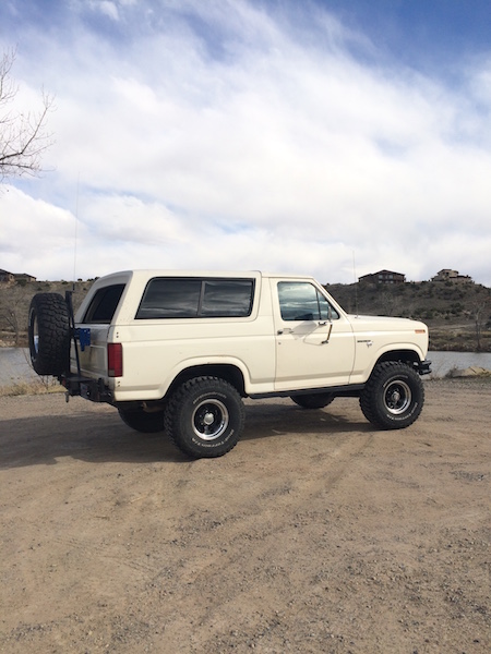 1981 Ford Bronco - A white 1980s Ford Bronco is parked on a dirt lot with a spare tire mounted on the rear. The SUV has tinted windows and is facing to the ri…