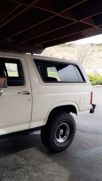 1981 Ford Bronco - Side view of a white SUV parked under a wooden structure, showing its rear quarter panel and off-road tires.