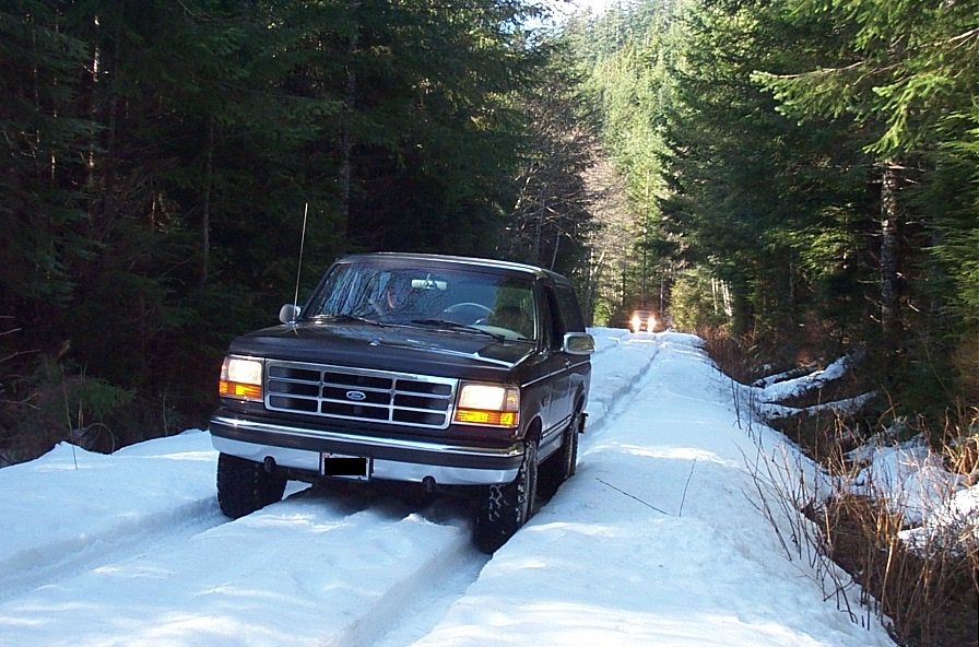 Dark Ford Bronco driving on a snowy forest road. Another vehicle is visible ahead