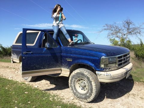 1996 Ford Bronco - A woman stands on the roof of a blue 1996 Ford Bronco, taking a photo with a camera.