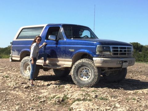 1996 Ford Bronco - A woman stands by a lifted blue Ford Bronco on a dry, rocky terrain under a clear blue sky.
