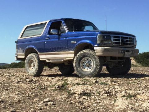 1996 Ford Bronco - A blue 1996 Ford Bronco with a white top is shown off-road on a sunny day.