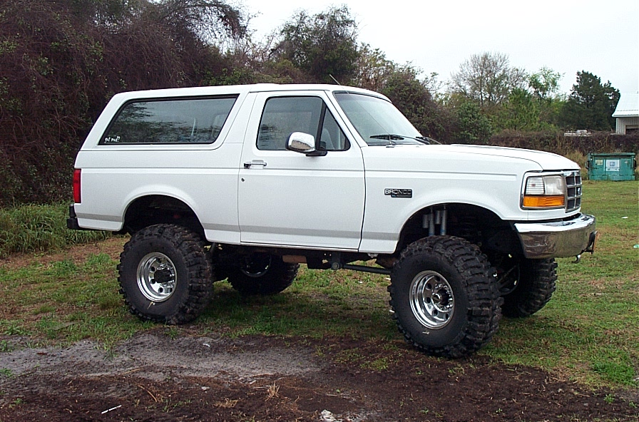 Lifted white two-door Ford Bronco on mud tires and chrome wheels in a grassy yard