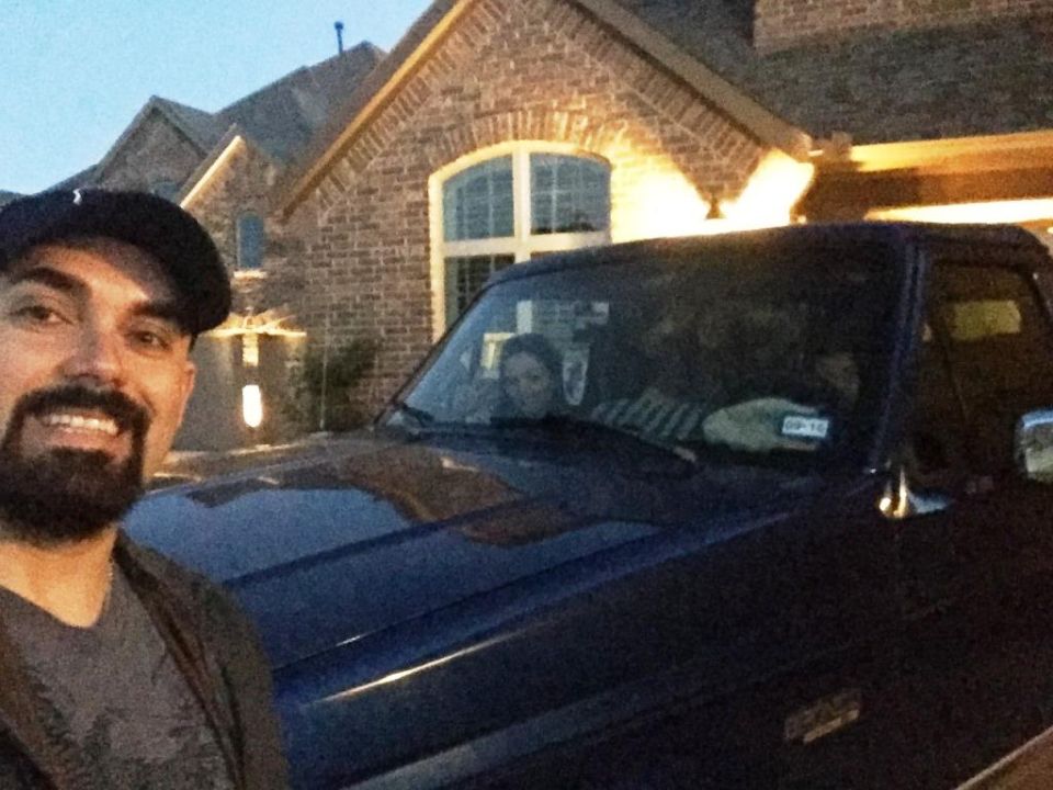 Man in baseball cap and beard smiles for selfie in front of a dark blue 1996 Ford Bronco. House in background