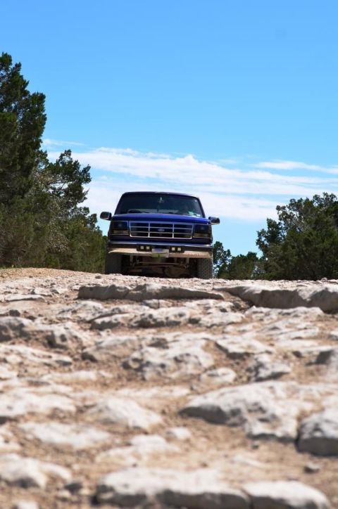 1996 Ford Bronco - A blue Ford F-Series truck navigates a rocky, off-road path under a bright blue sky.
