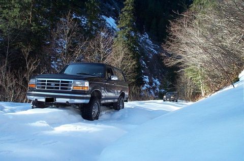 1995 Ford Bronco - A dark brown Ford Bronco navigates a snowy mountain road, with another vehicle following behind.