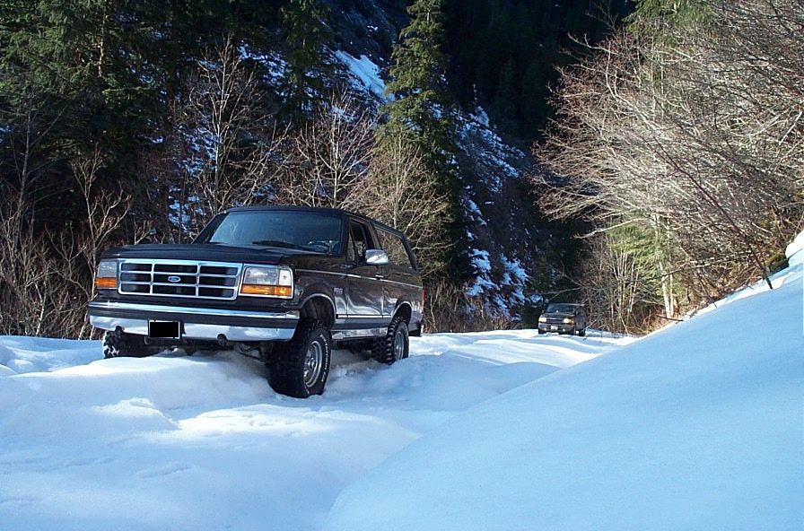 Dark brown Ford Bronco driving on a snow-covered road, front 3/4 view. Another vehicle is visible in the distance