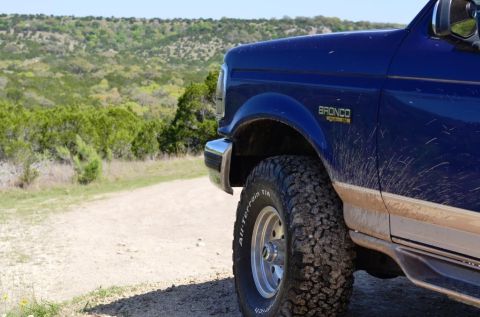 1996 Ford Bronco - A blue 1996 Ford Bronco is seen from the front quarter angle on a dirt road with a scenic, hilly background.