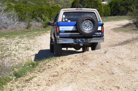 1996 Ford Bronco - This two-tone Ford Bronco is driving on a dusty, off-road trail with brush and trees in the background.