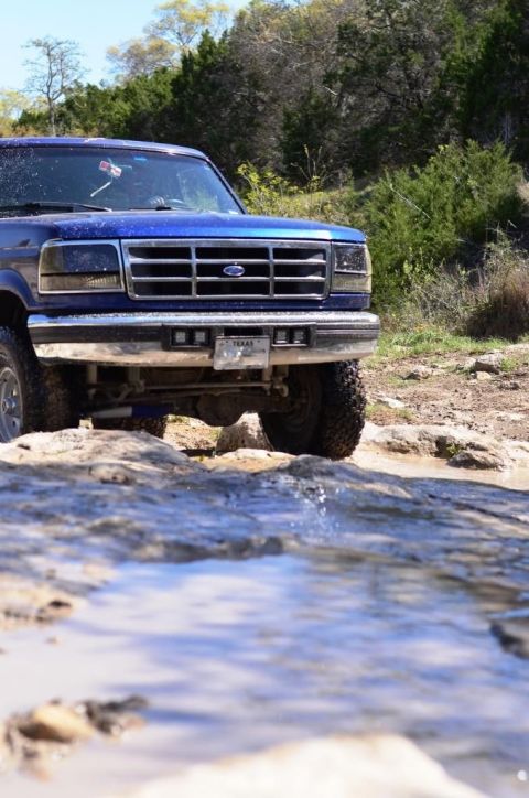 1996 Ford Bronco - A blue Ford truck navigates a rocky, wet terrain under a clear sky.