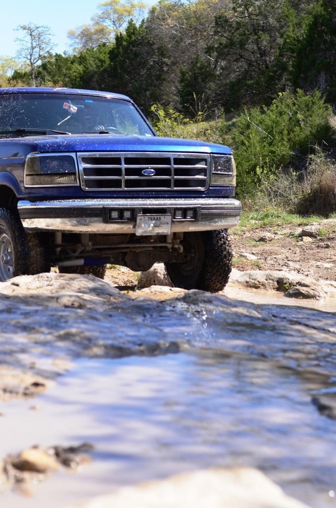 Blue Ford truck driving over rocks and water. Texas plate visible