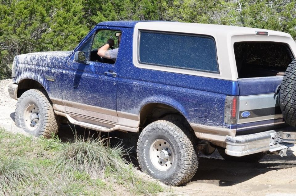Mud-splattered blue Ford Bronco with a white top, seen from the rear three-quarter angle on a dirt path