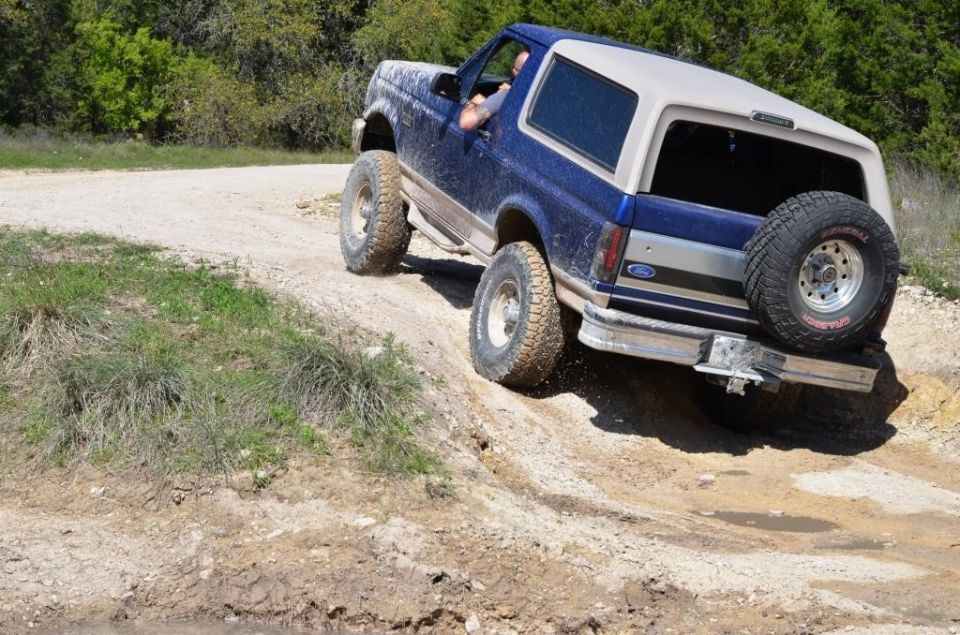 Blue and tan Ford Bronco off-roading on a dirt path, rear-mounted spare tire visible