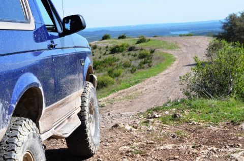 1996 Ford Bronco - A blue SUV with off-road tires navigates a dusty, winding dirt road with a scenic overlook in the distance.