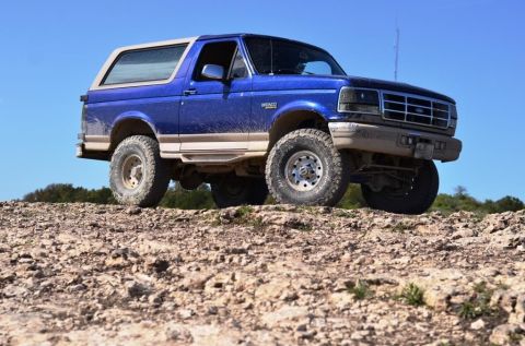 1996 Ford Bronco - This blue Ford Bronco is shown off-roading on a rocky terrain with a clear sky above.