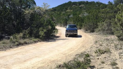 1996 Ford Bronco - A blue 1996 Ford Bronco navigates a dirt road in a hilly, wooded landscape.