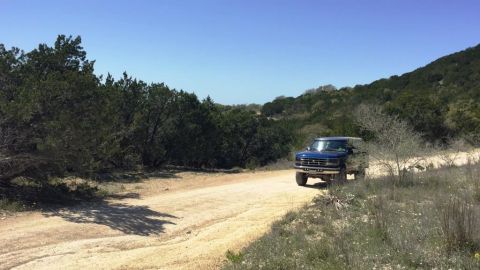 1996 Ford Bronco - A blue pickup truck navigates a dusty, unpaved road surrounded by trees and hills under a clear blue sky.