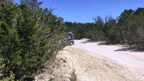 1996 Ford Bronco - An SUV navigates a dusty off-road trail surrounded by dense vegetation under a clear blue sky.