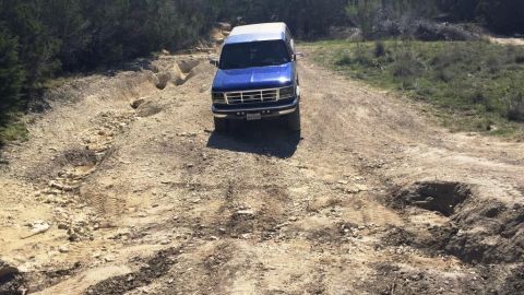 1996 Ford Bronco - A blue SUV navigates a challenging dirt path with significant ruts and uneven terrain.
