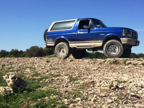 1996 Ford Bronco - A blue 1996 Ford Bronco navigates a , rocky landscape under a bright blue sky.