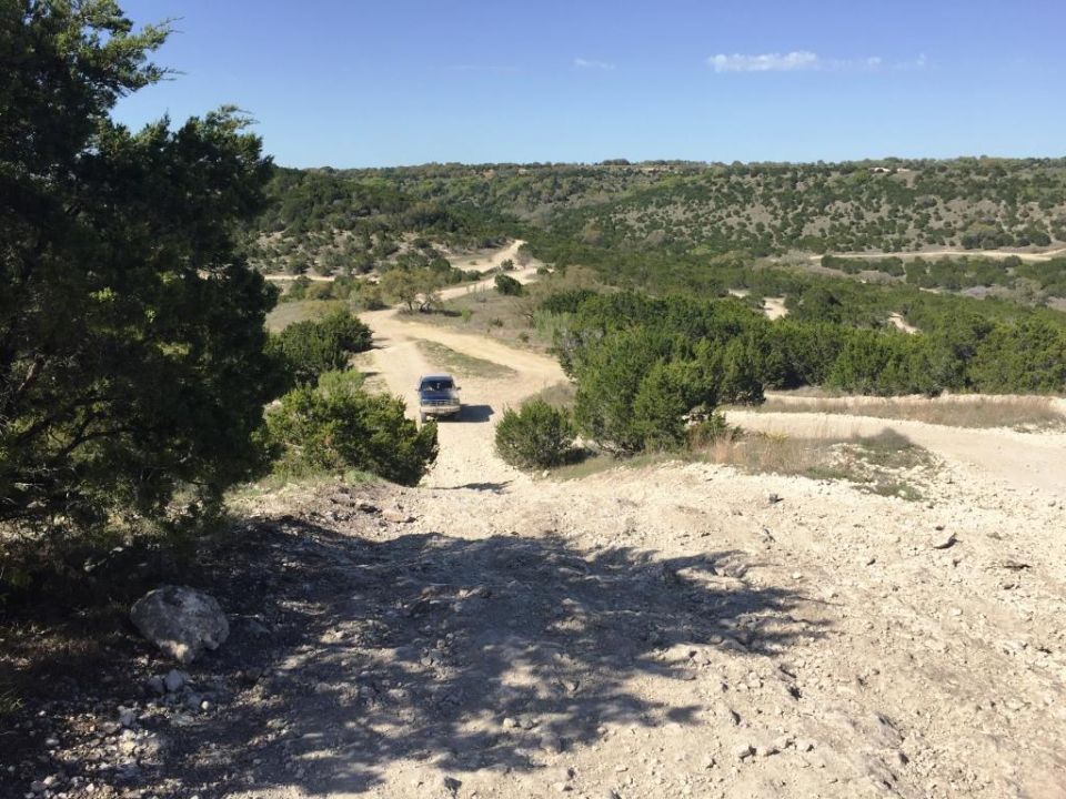 Dark SUV drives on a winding dirt road through a dry, hilly landscape with scattered trees