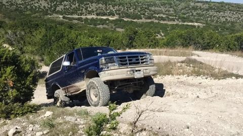 1996 Ford Bronco - A blue 1996 Ford Bronco tackles an off-road trail, kicking up dust as it climbs.