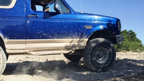 1996 Ford Bronco - This blue Ford Bronco is shown off-roading on a dirt path, with mud splattered on its side.