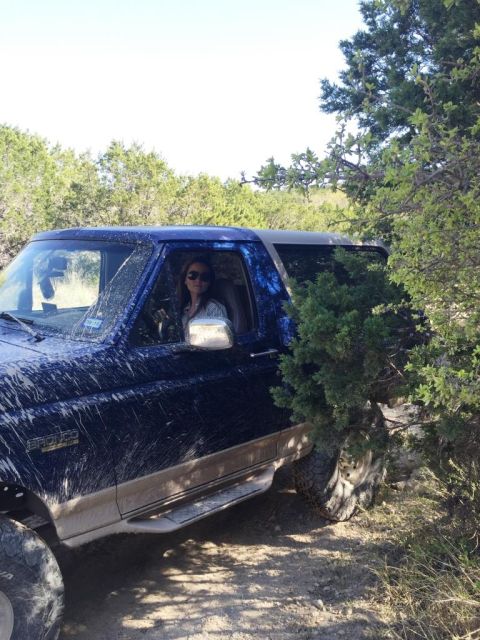 1996 Ford Bronco - A woman drives a muddy blue 1996 Ford Bronco through a wooded, off-road area.