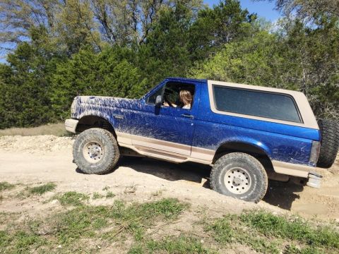 1996 Ford Bronco - A blue Ford Bronco navigates a dirt trail, its tires kicking up mud.