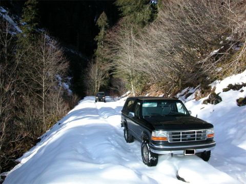 1995 Ford Bronco - Two SUVs navigate a snowy mountain pass in a wooded area during winter.