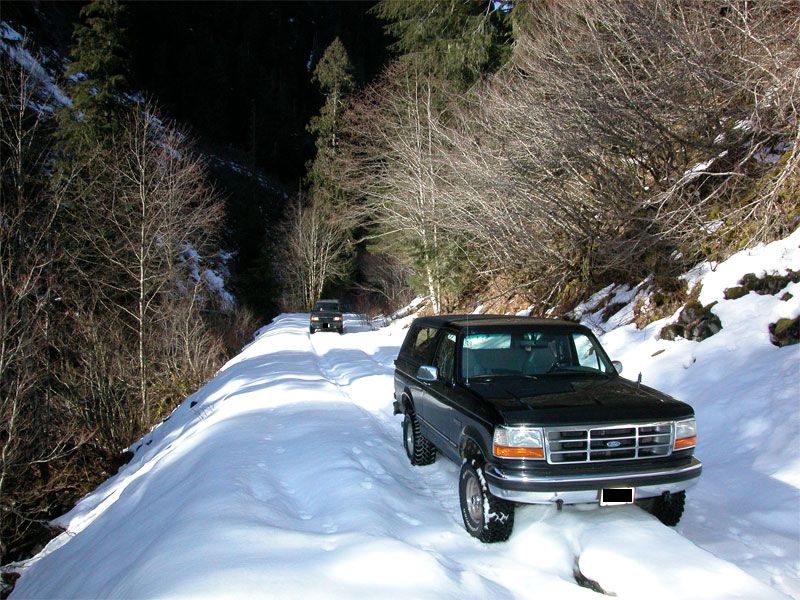 Black 1995 Ford Bronco driving on a snow-covered mountain road with another vehicle behind it