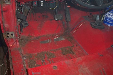 1974 Ford Bronco - The interior floor of a red vehicle shows signs of wear and tear, with dirt, rust, and exposed wiring visible.