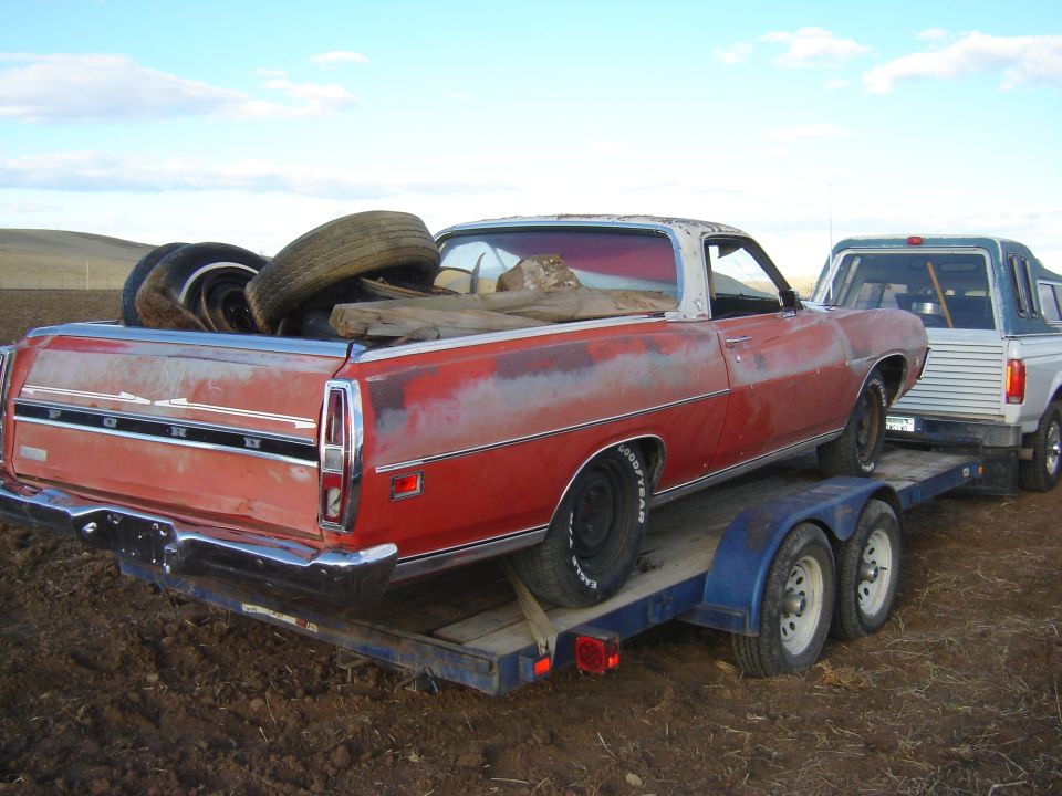 Red Ford Ranchero on a trailer, loaded with tires and wood. A white pickup truck is towing it