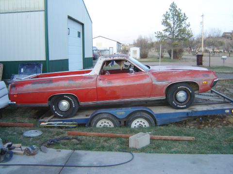 1971 Ford Ranchero - A red pickup truck with a white top is loaded onto a trailer, parked on grassy ground near a large building.