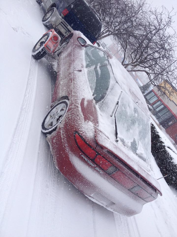 Red Ford Probe covered in snow, parked on a snowy street next to a blue SUV.