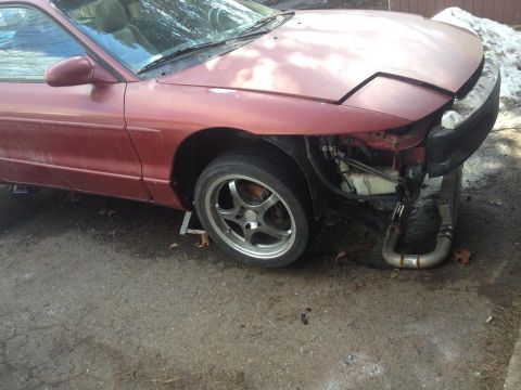 1995 Ford Probe - The front driver's side of a red Ford Probe is shown, jacked up on a stand. The front bumper is removed, revealing the engine components an…