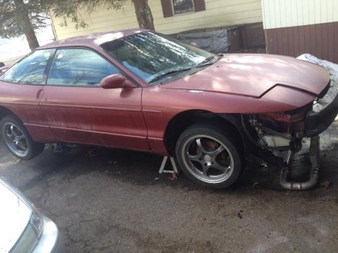 1995 Ford Probe - A red Ford Probe is jacked up on stands with its front bumper and headlight assembly removed, revealing the engine bay and exhaust system.