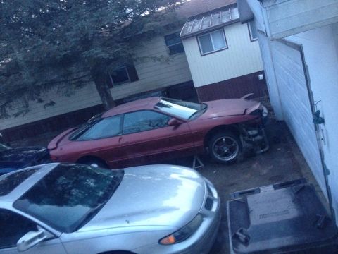 1995 Ford Probe - A red 1995 Ford Probe is parked next to a silver car and a garage. The red car's front end is damaged and appears to be missing body panels.