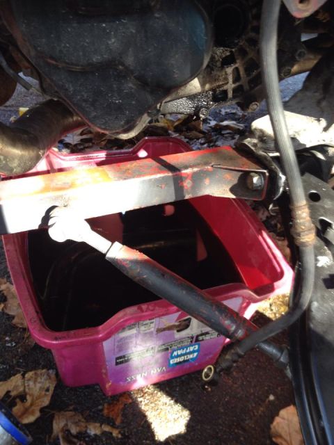 1995 Ford Probe - A red and black oil drain pan is positioned beneath a vehicle's engine, collecting dark fluid. The pan has text that includes "VAN NESS ENC…