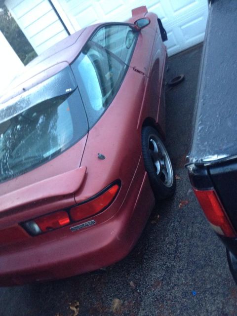1995 Ford Probe - The rear passenger side of a red 1995 Ford Probe is shown parked on asphalt in front of a garage. The car has a spoiler and a visible exhau…