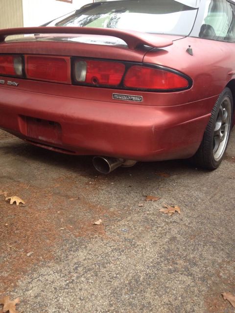 1995 Ford Probe - The rear driver's side of a red coupe is shown, featuring a spoiler, taillights, and a visible exhaust pipe. The car is parked on a gravel…