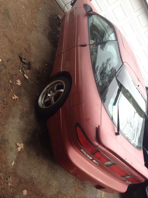1995 Ford Probe - The rear passenger side of a red coupe is shown parked on a dirt and concrete surface next to a garage door. The car has a spoiler and a vi…