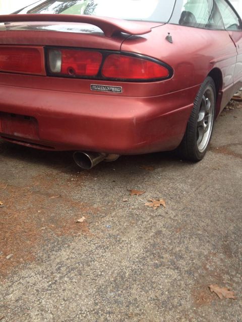 1995 Ford Probe - The rear passenger side of a red MazdaSpeed branded Ford Probe is shown parked on a concrete surface with scattered leaves and pine needles…