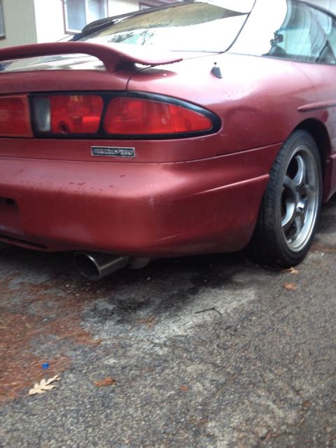 1995 Ford Probe - The rear passenger side of a red Ford Probe is shown parked on an asphalt surface with scattered pine needles. A chrome exhaust tip is visi…