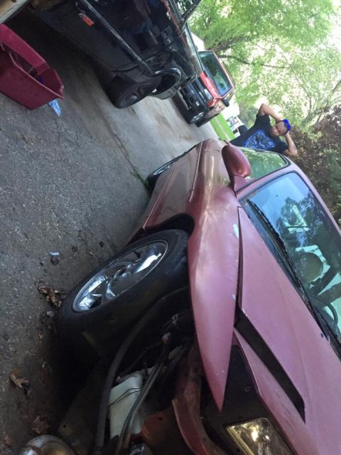 1995 Ford Probe - A damaged maroon car is parked on a driveway next to a black pickup truck. A person is lying on the ground behind the pickup truck.