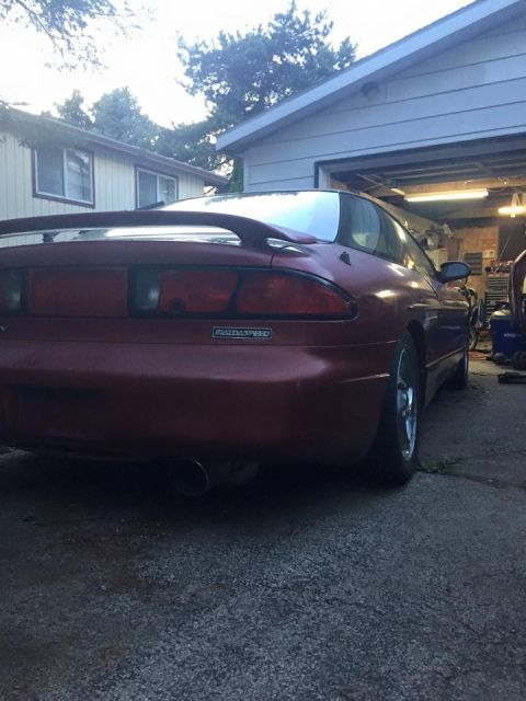 1995 Ford Probe - The rear passenger side of a dark red MazdaSpeed coupe is shown parked on a gravel driveway in front of an open garage. A spoiler is visibl…