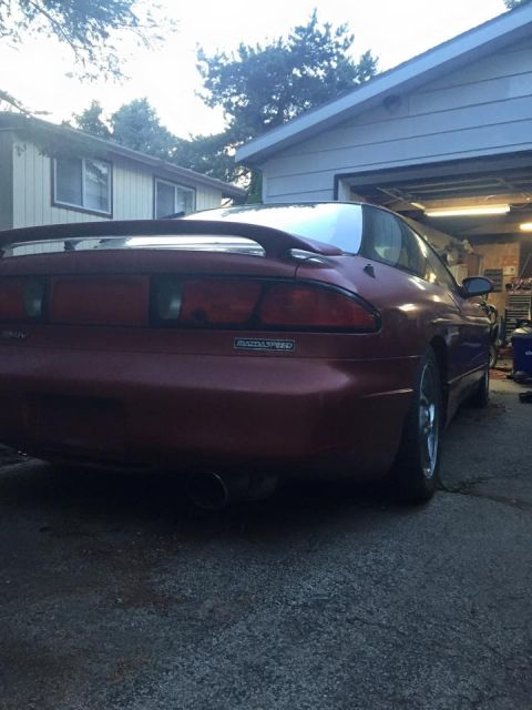 1995 Ford Probe - The rear driver's side quarter view of a red MazdaSpeed branded coupe is shown parked on a driveway in front of a garage. The car features…