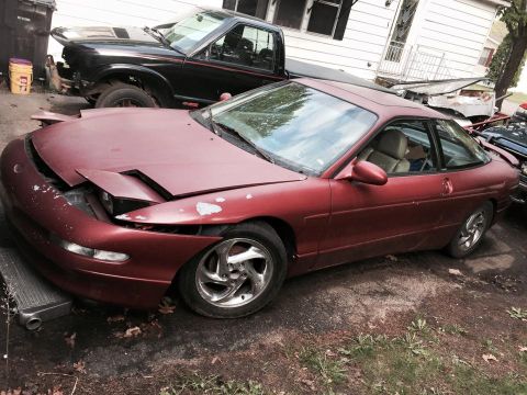 1995 Ford Probe - A maroon Ford Probe is parked outdoors next to a black pickup truck. The Probe's hood is open and its paint is chipped.