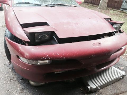 1995 Ford Probe - The front of a red, slightly faded Ford Probe is shown, with its pop-up headlight partially raised. An intercooler is visible underneath th…